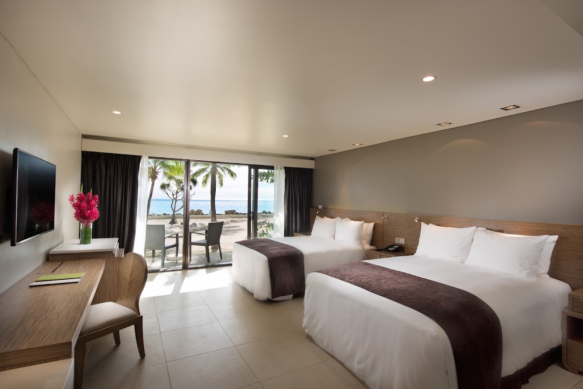 Resort guest room with two queen beds, wooden desk, flat-screen TV, and glass doors opening to a beachfront terrace with palm trees and ocean view.