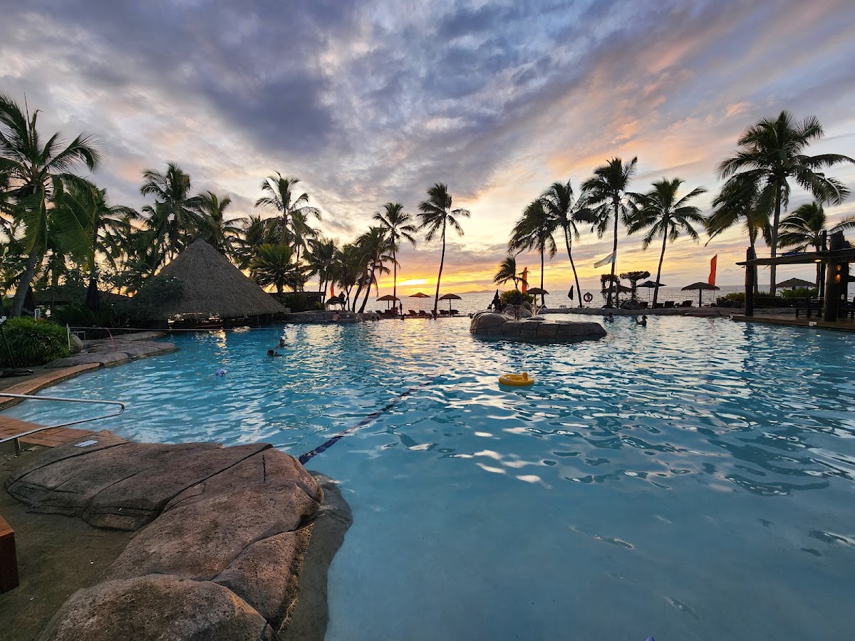 Resort pool at sunset with palm trees, thatched-roof bar, and ocean horizon under colorful clouds.
