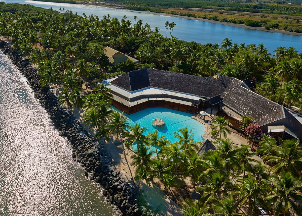 Aerial view of a curved resort building with turquoise pool surrounded by palm trees on a rocky shoreline, with a wide river and lush countryside beyond.