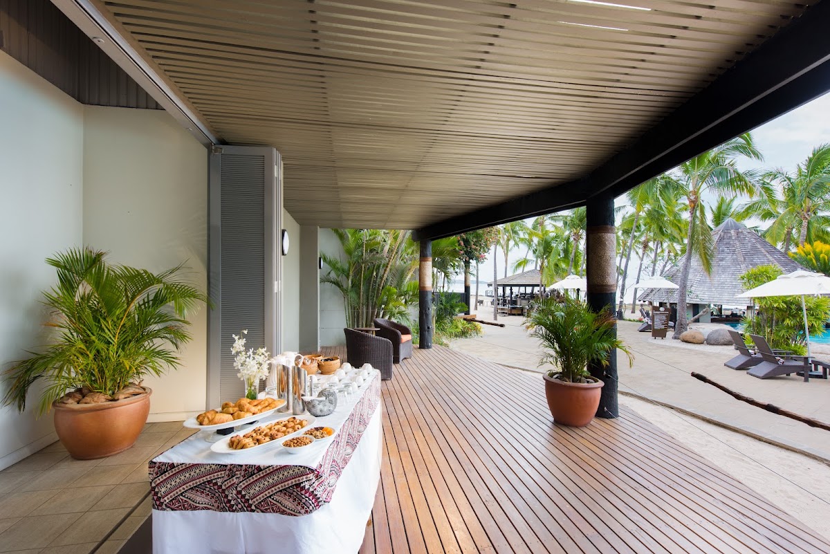 Covered beachfront terrace with food service table, wooden deck, palm trees, and pool area with umbrellas visible in the distance.