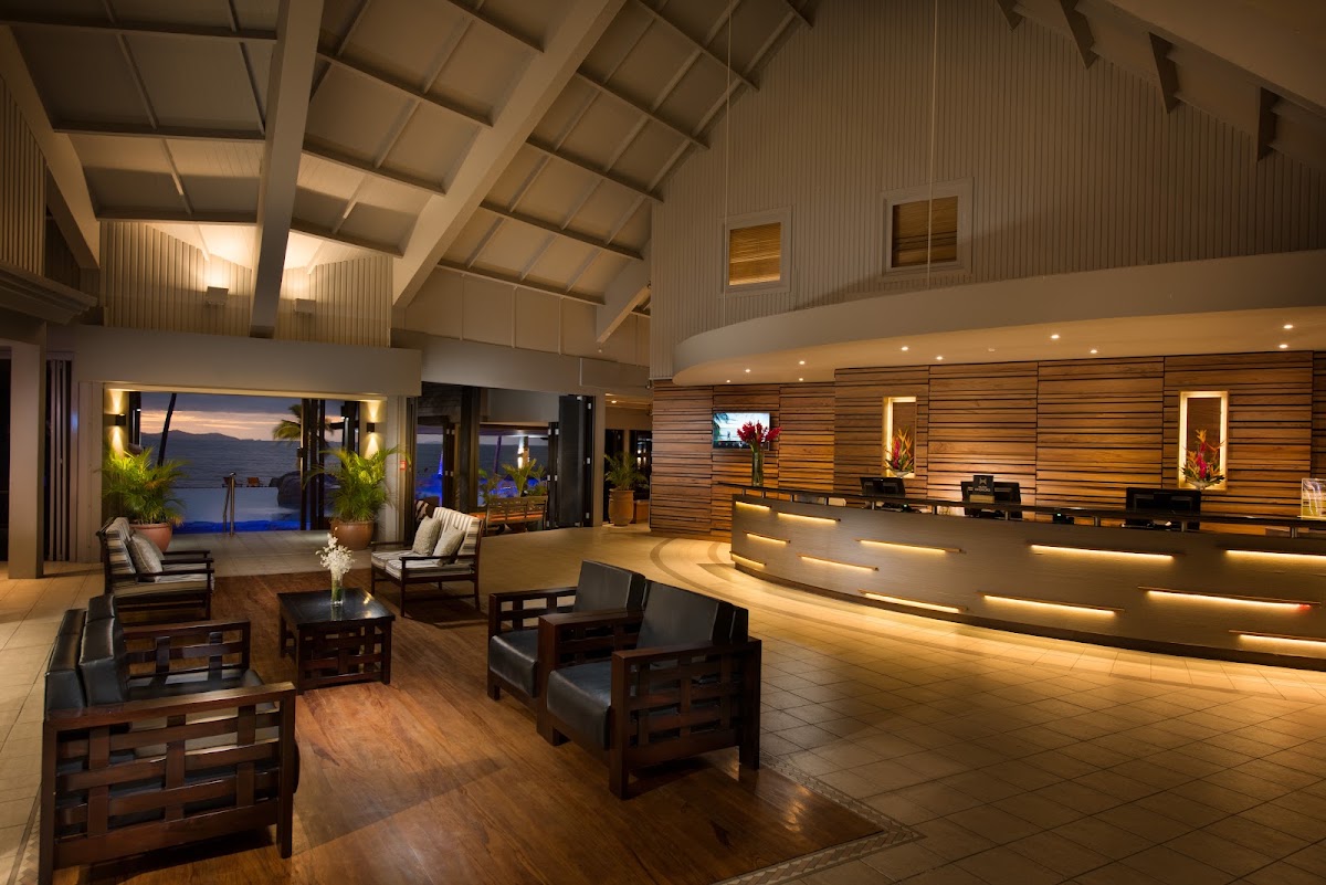 Modern resort lobby with wooden furniture, reception desk, and ocean view through floor-to-ceiling windows at dusk.