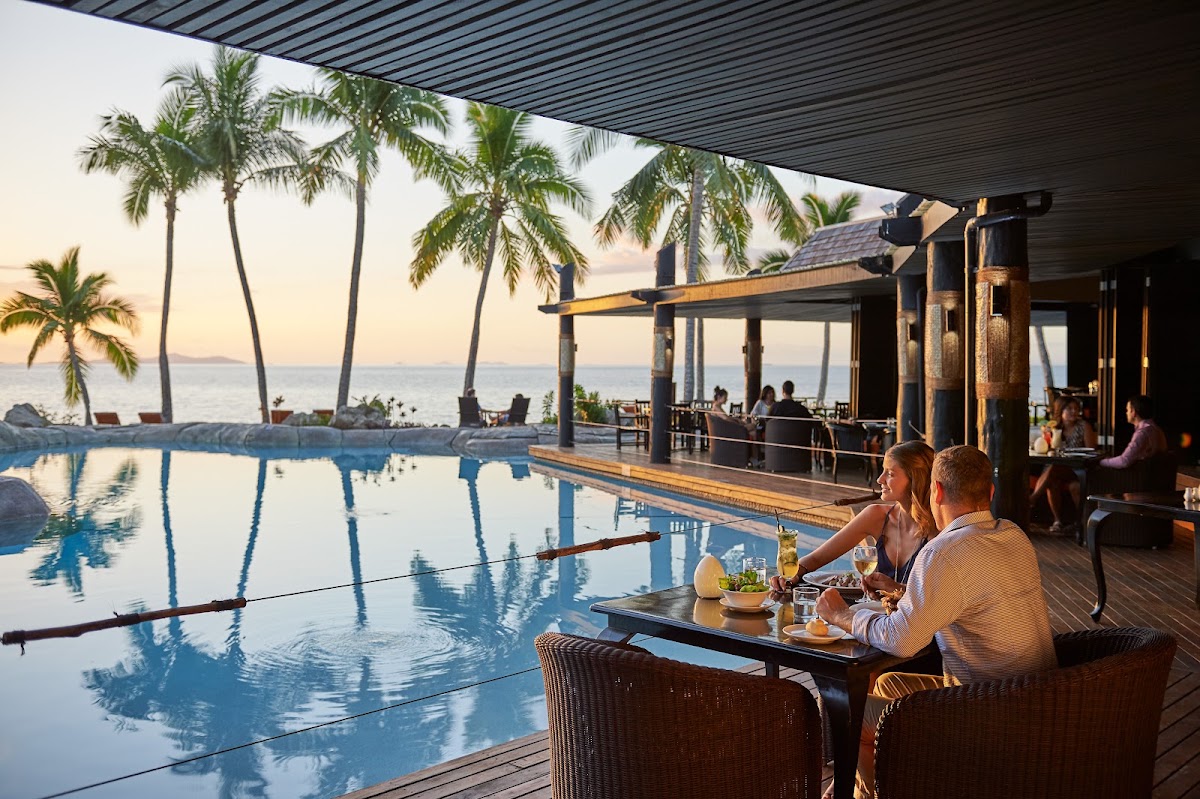 Guests dine poolside at sunset with palm trees and ocean views beyond the infinity pool at DoubleTree Resort by Hilton Hotel Fiji.