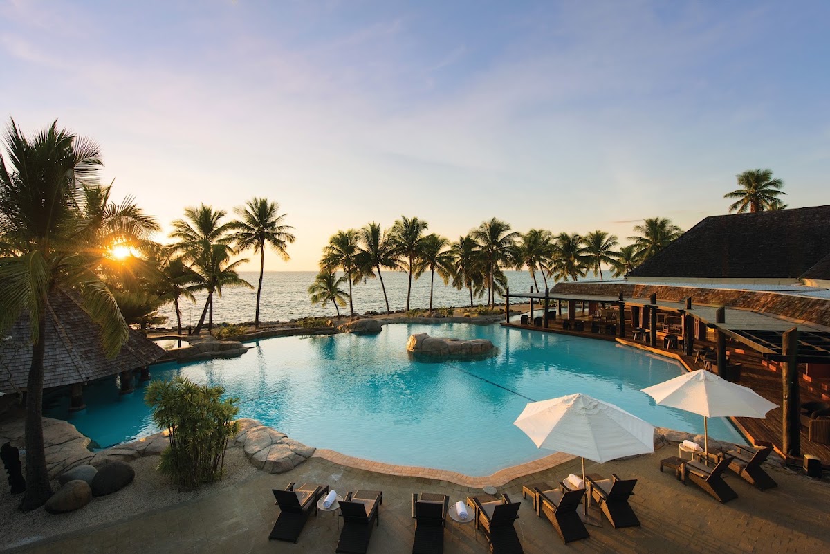 Curved pool with turquoise water at sunset, surrounded by palm trees, lounge chairs with white umbrellas, and ocean view beyond at DoubleTree Resort Fiji Sonaisali Island.