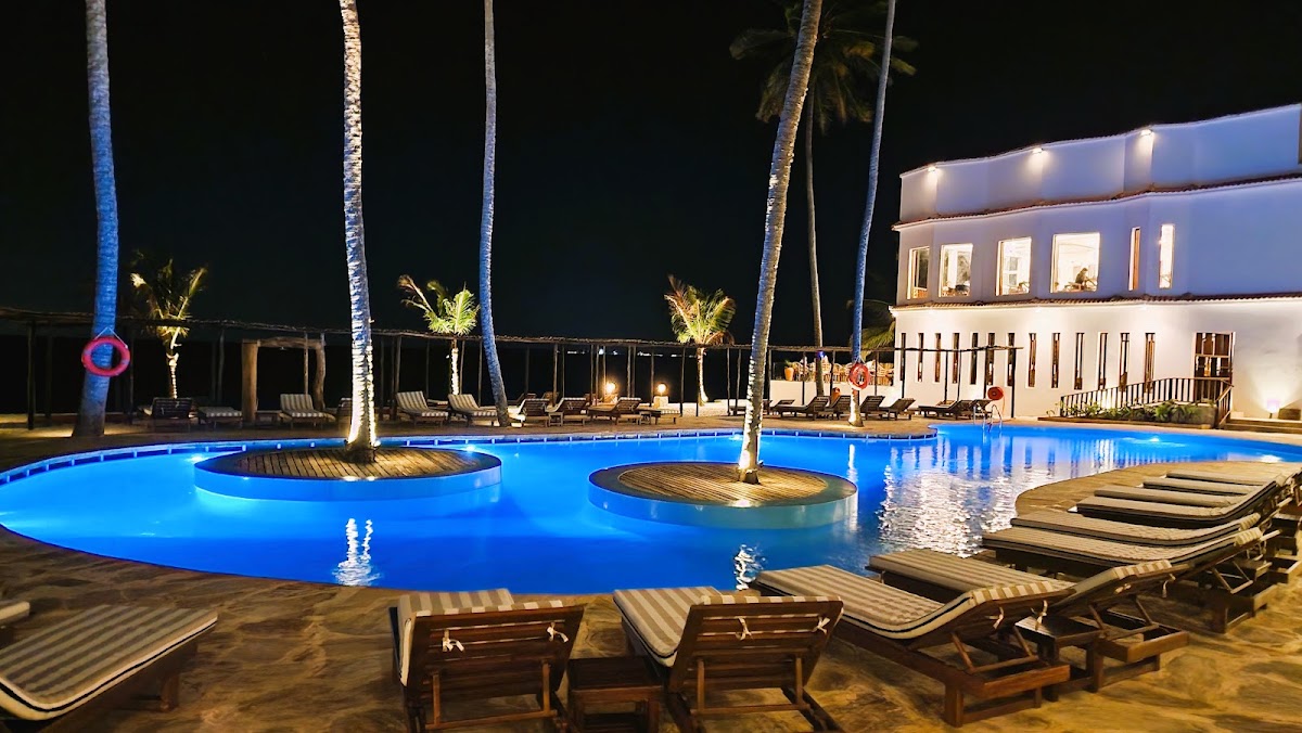 Lit blue pool with palm trees at night, striped lounge chairs in foreground, white resort building and ocean view beyond.