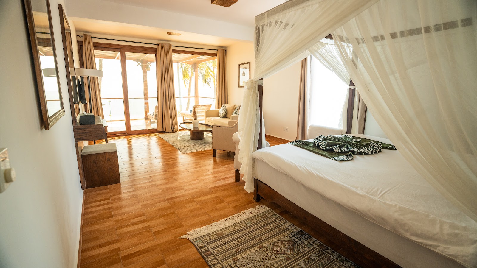 Four-poster bed with white sheer canopy in spacious resort suite with wooden floors, ocean view balcony, and sitting area.