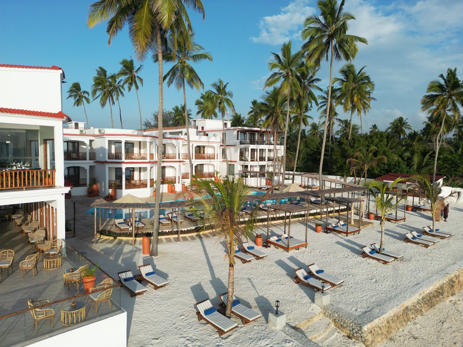 White and red resort buildings with palm trees overlook a sandy beach dotted with blue loungers and thatched umbrellas at Dream of Zanzibar Resort & Spa.