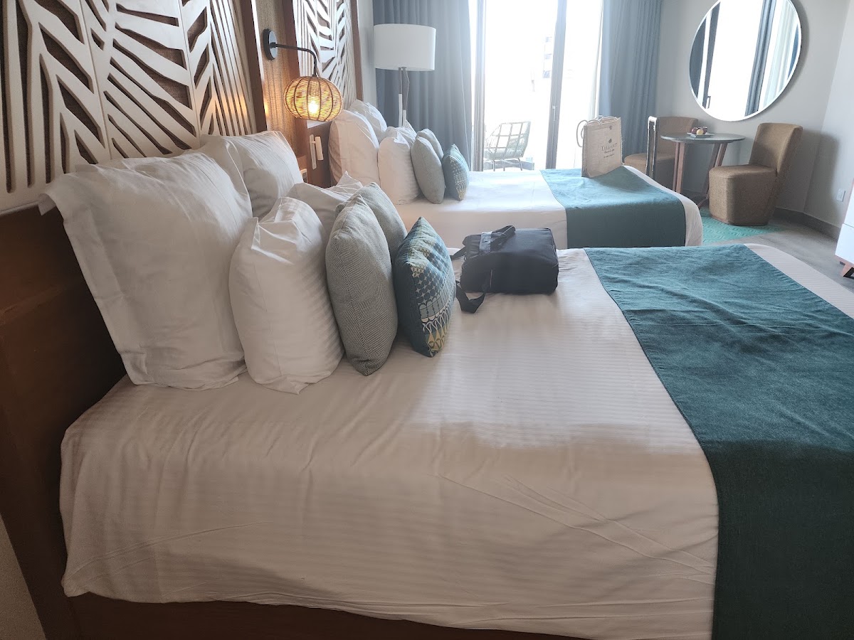 Resort bedroom with white pillows arranged on a bed with teal throw blanket, pendant lighting, and ocean-view balcony doors.