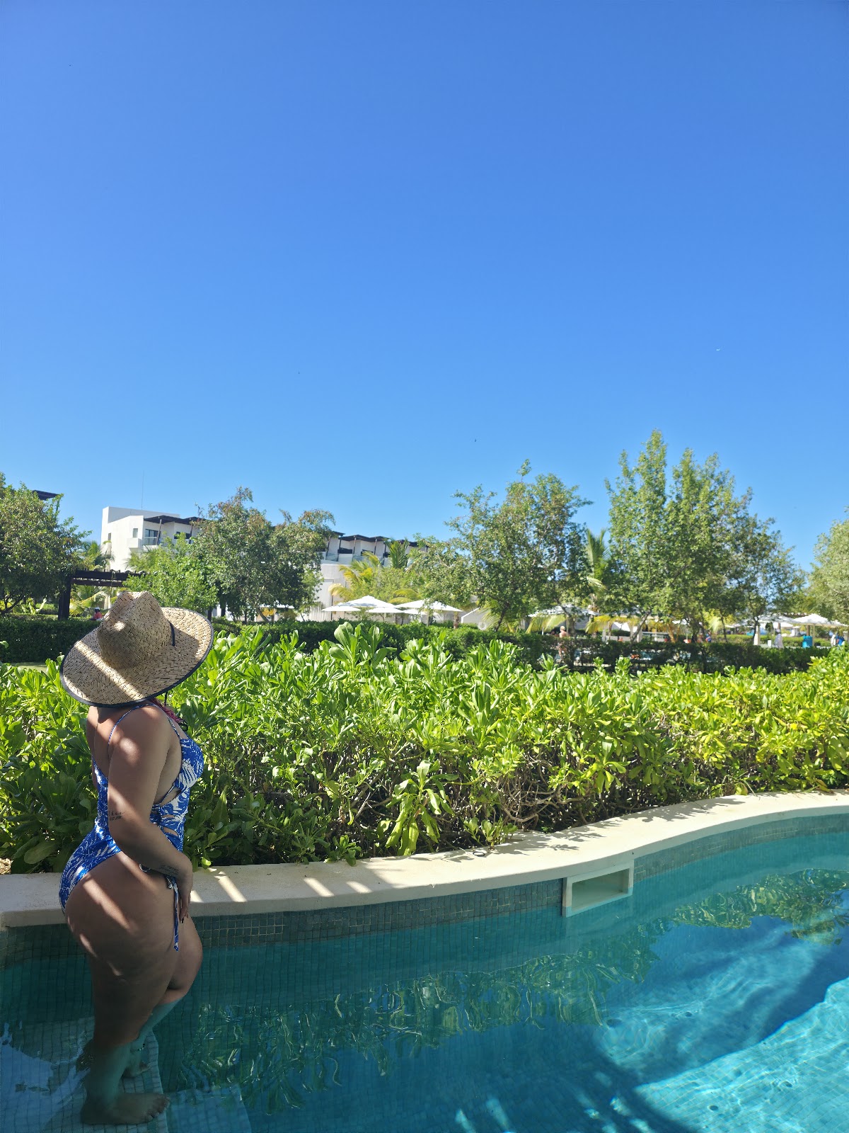 Woman in blue swimsuit and straw hat stands by resort pool overlooking mangrove wetlands and white buildings under clear sky.