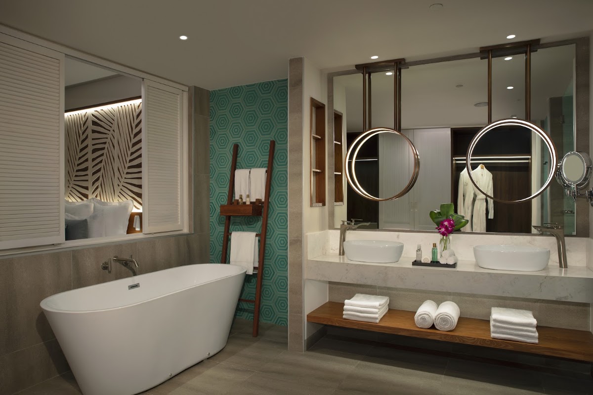 Bathroom with freestanding white tub, double vanity with circular mirrors, turquoise patterned accent wall, and wooden shelf holding rolled towels.