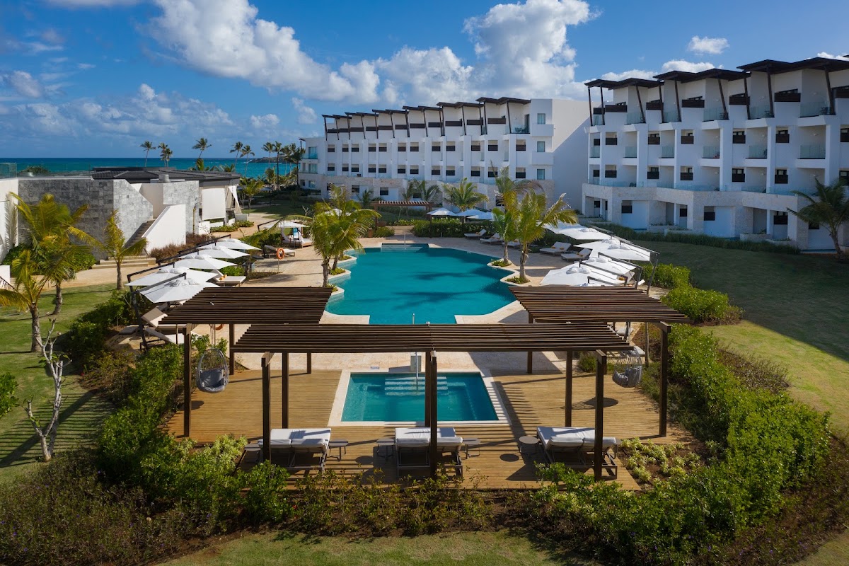 Aerial view of Dreams Macao Beach Punta Cana with two turquoise pools, white loungers, black pergolas, palm trees, and ocean beyond white resort buildings.