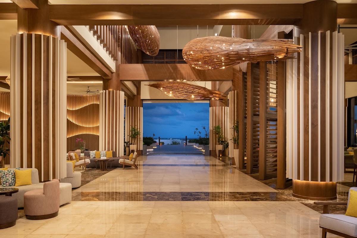 Modern lobby with polished marble floors, wooden sculptural fish overhead, and open view to twilight beach and ocean beyond.
