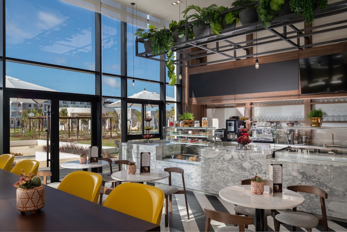 Modern cafe interior with yellow chairs, marble counters, hanging plants, and floor-to-ceiling windows overlooking the resort grounds and beach beyond.