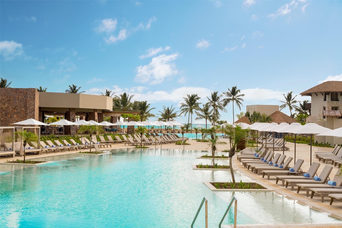 Large turquoise pool with lounge chairs and white umbrellas at Dreams Playa Esmeralda, palm trees and beige buildings visible beyond.