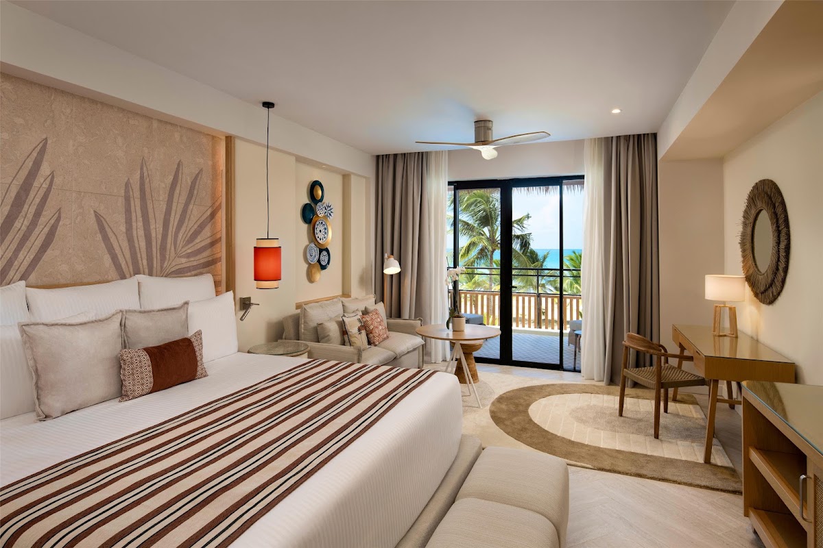 Resort room with king bed, seating area, and ocean view balcony with palm trees visible through glass doors.