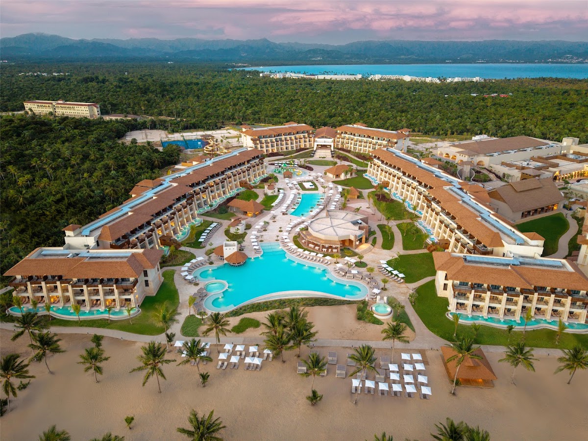 Aerial view of Dreams Playa Esmeralda Resort & Spa with curved hotel buildings, large pool complex, and beach with lounge chairs backed by tropical forest and mountains.
