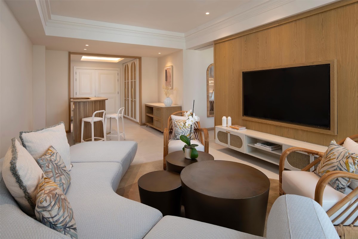 Resort suite living room with light blue seating, round black tables, wall-mounted TV, wood paneling, and entry to bedroom area.