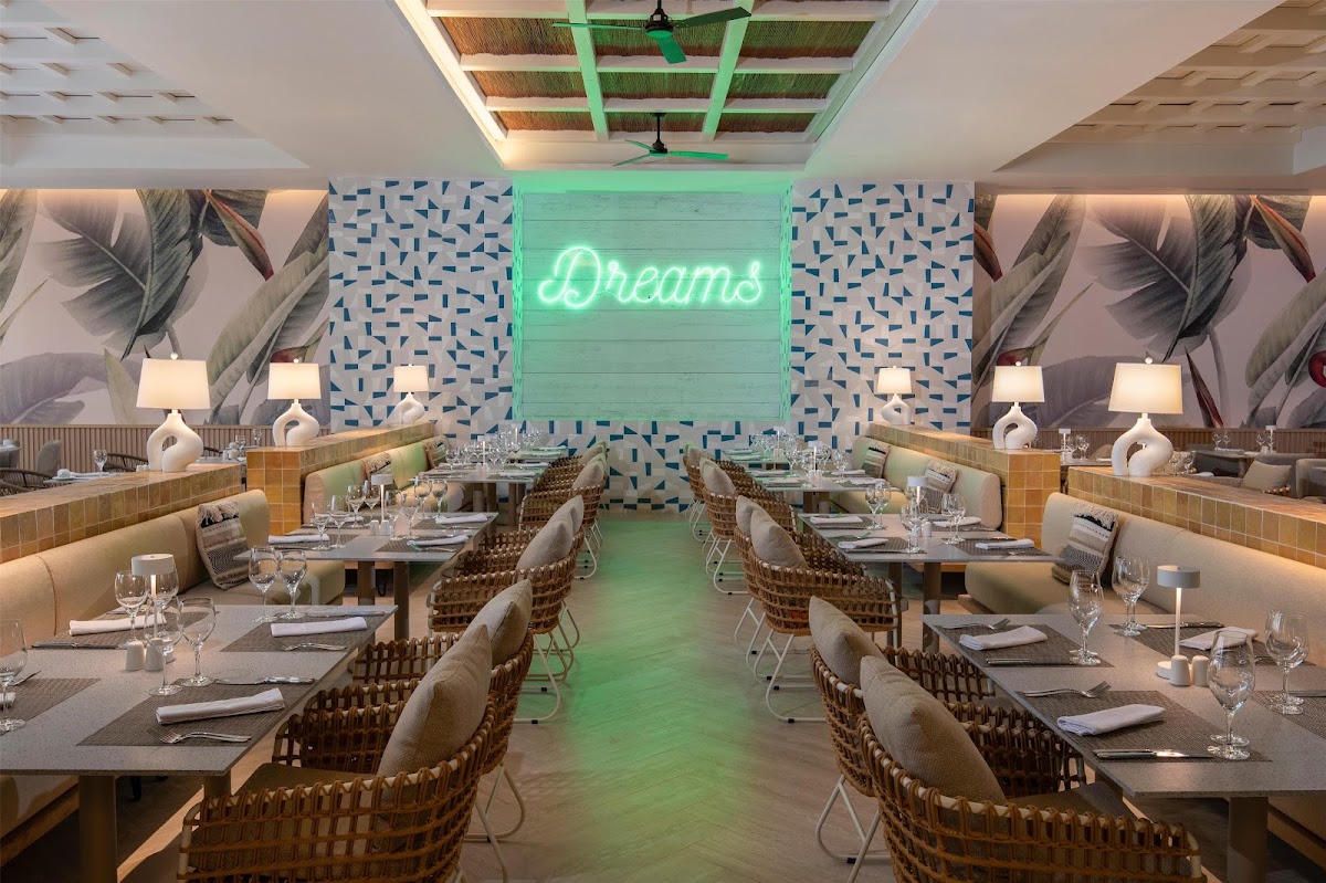 Modern restaurant dining room with woven chairs at tables, neon Dreams sign on patterned blue wall, and palm leaf murals.