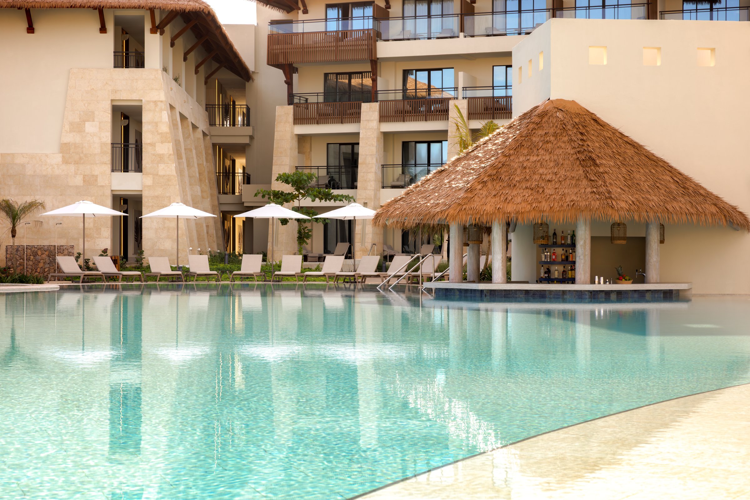 Resort pool with a thatched-roof swim-up bar, loungers and umbrellas below the guest buildings.