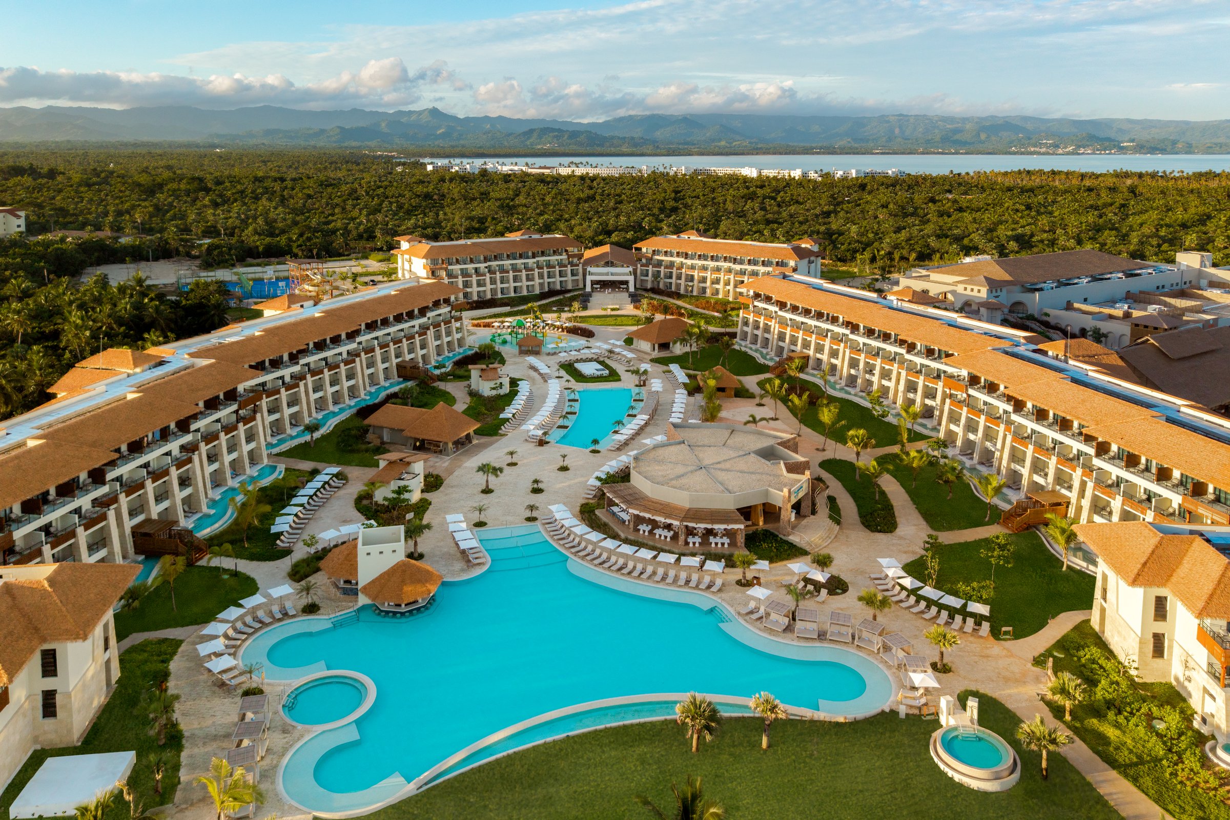 Wide aerial of the resort wings around the main pool complex, with forest and mountains beyond.