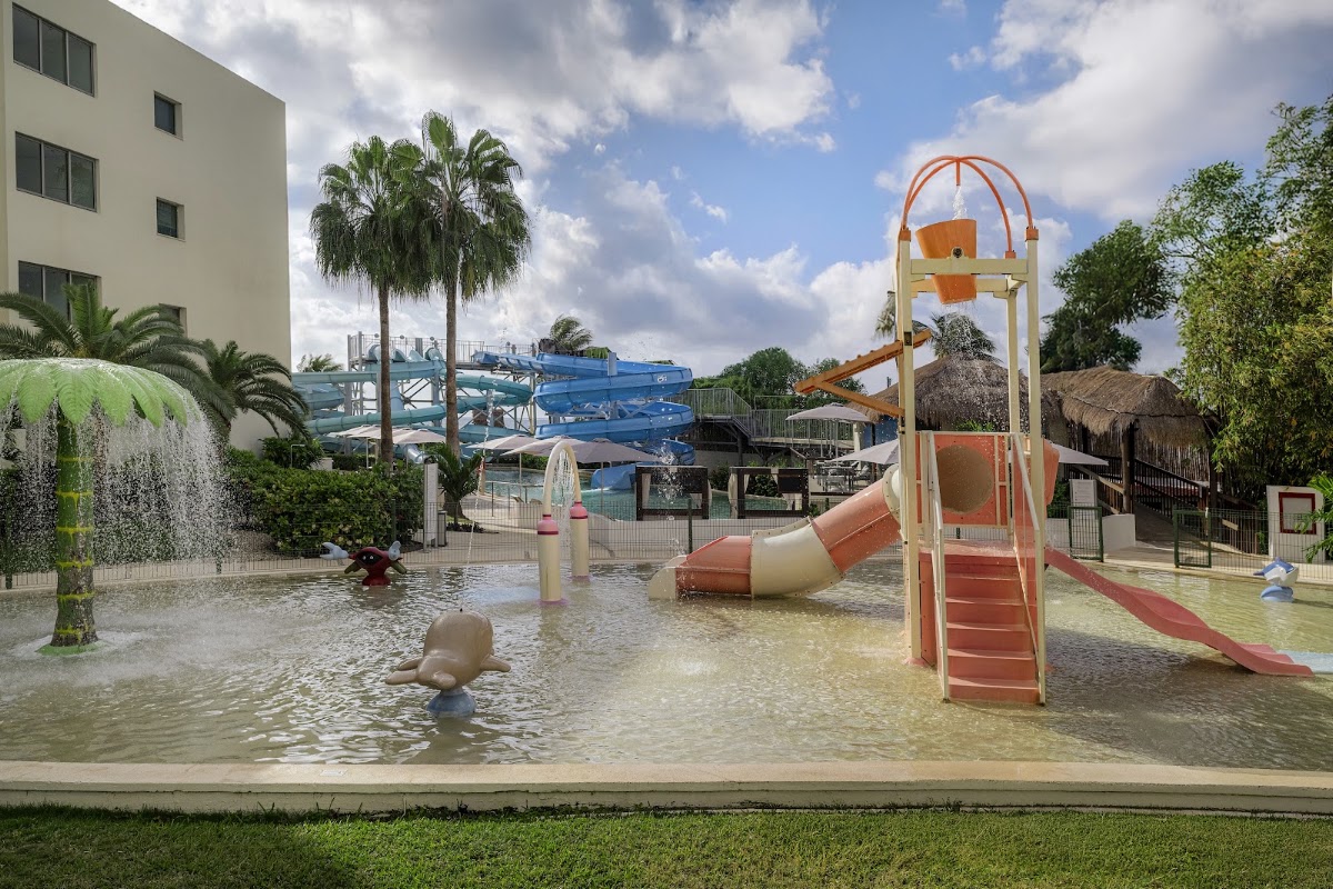 Children play in a shallow water park area with slides and a climbing structure, palm trees, and blue water slides visible behind a multi-story resort building.