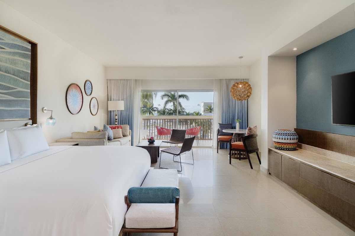 Spacious resort bedroom with white bedding, balcony overlooking palm trees, sitting area with red chairs, and ocean view beyond the railing.