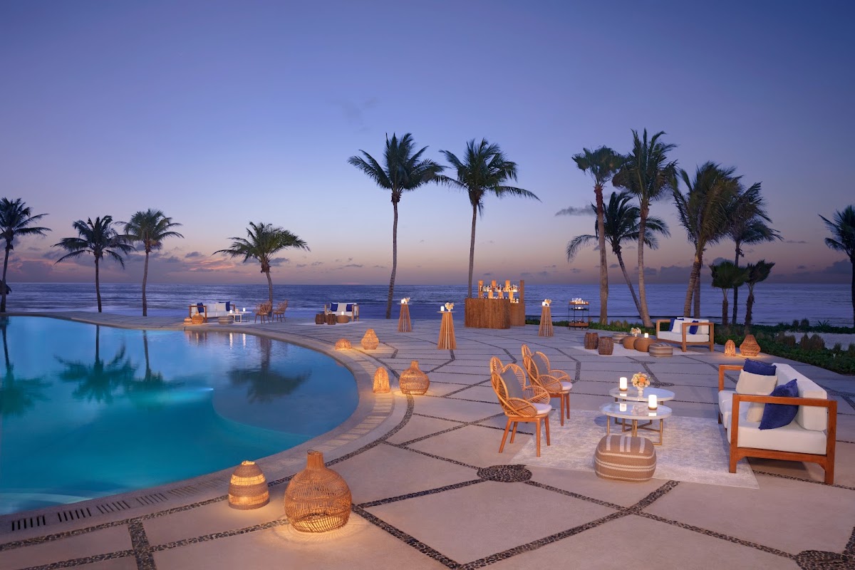 Beachfront pool deck at dusk with illuminated lanterns, lounge chairs, and palm trees overlooking the ocean at Dreams Puerto Morelos Resort & Spa.