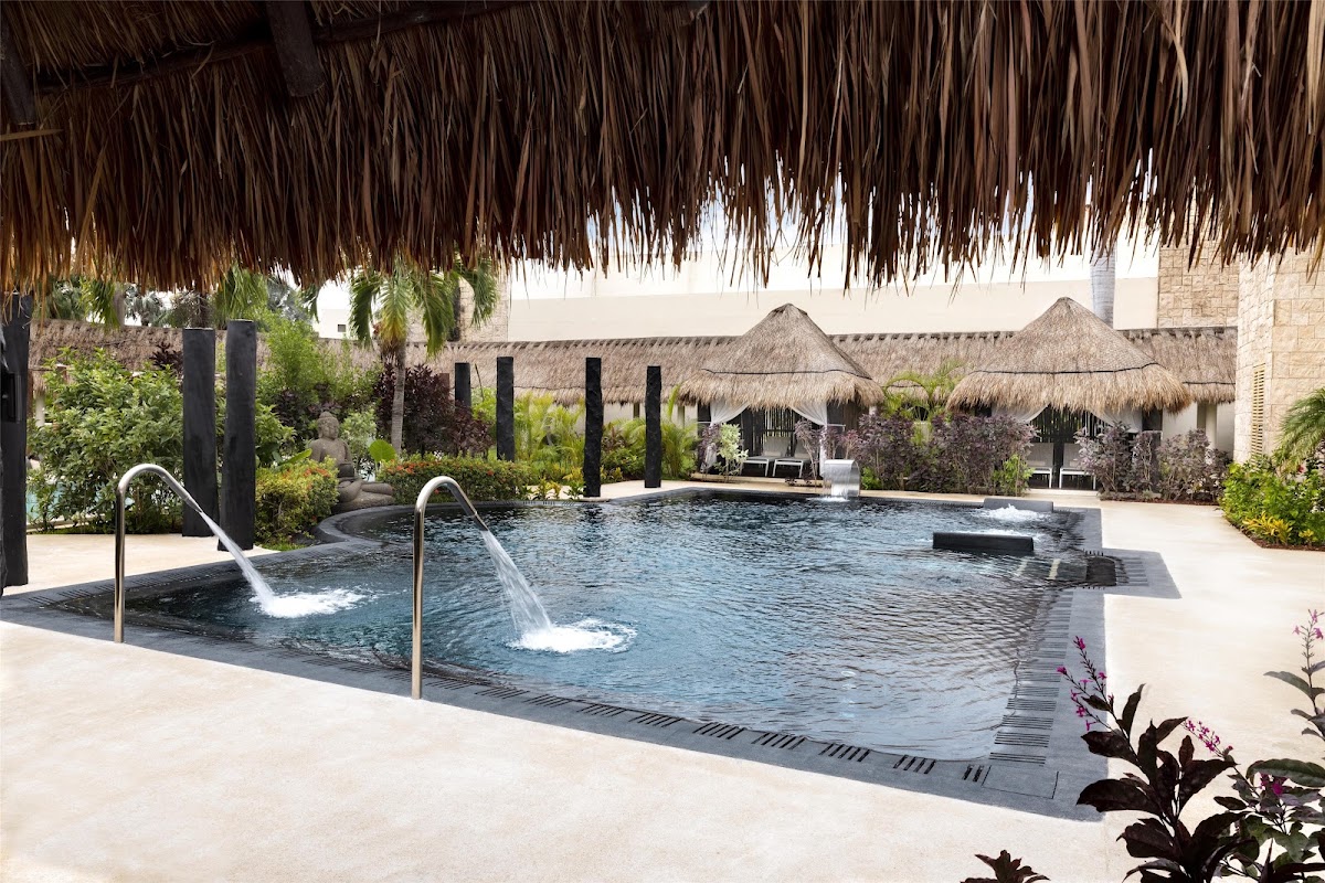 Resort pool with two waterfall features, thatched-roof palapas, black columns, and tropical landscaping along pale stone decking.