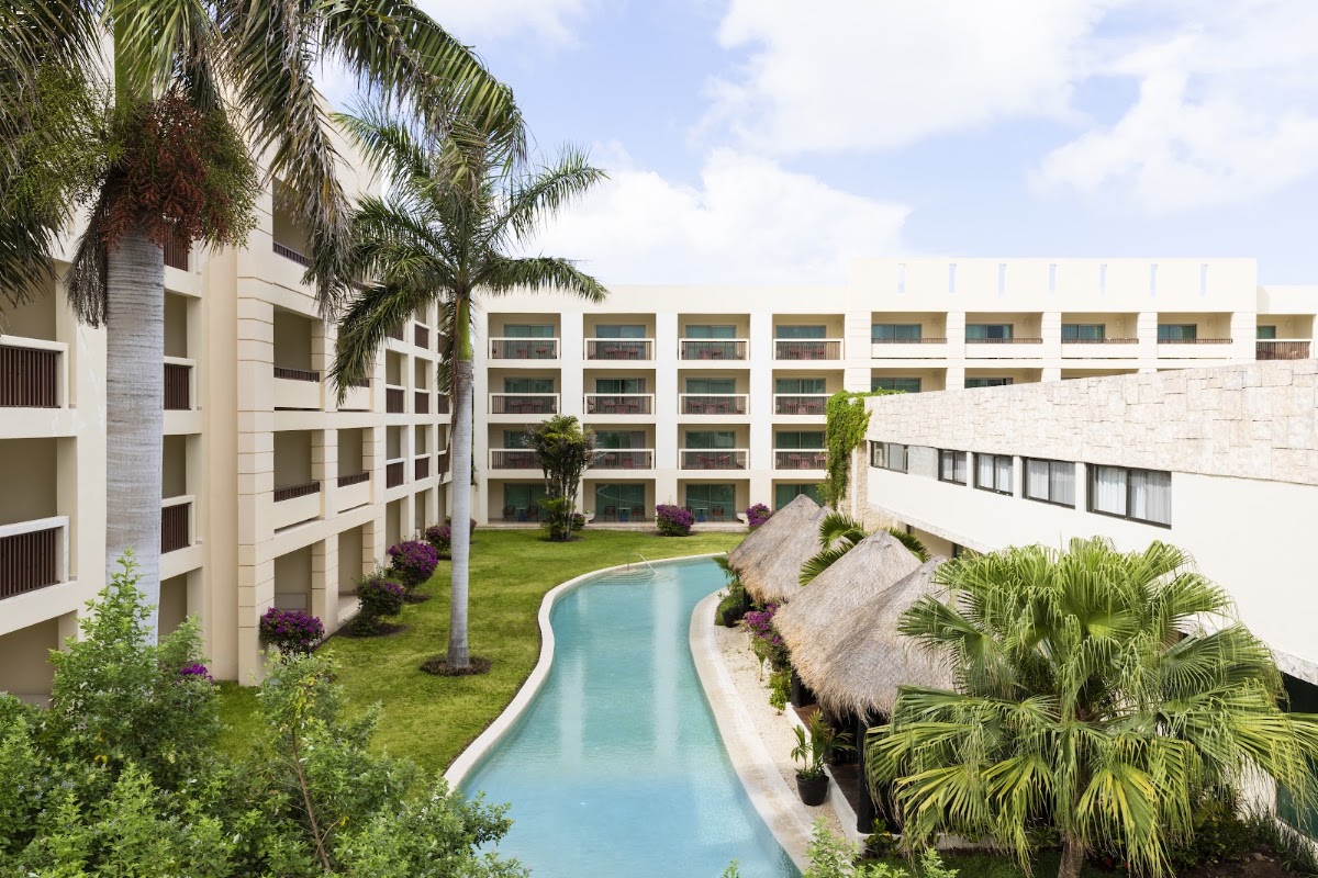 Curved turquoise pool winds through manicured courtyard surrounded by cream-colored resort buildings with balconies and palm trees.