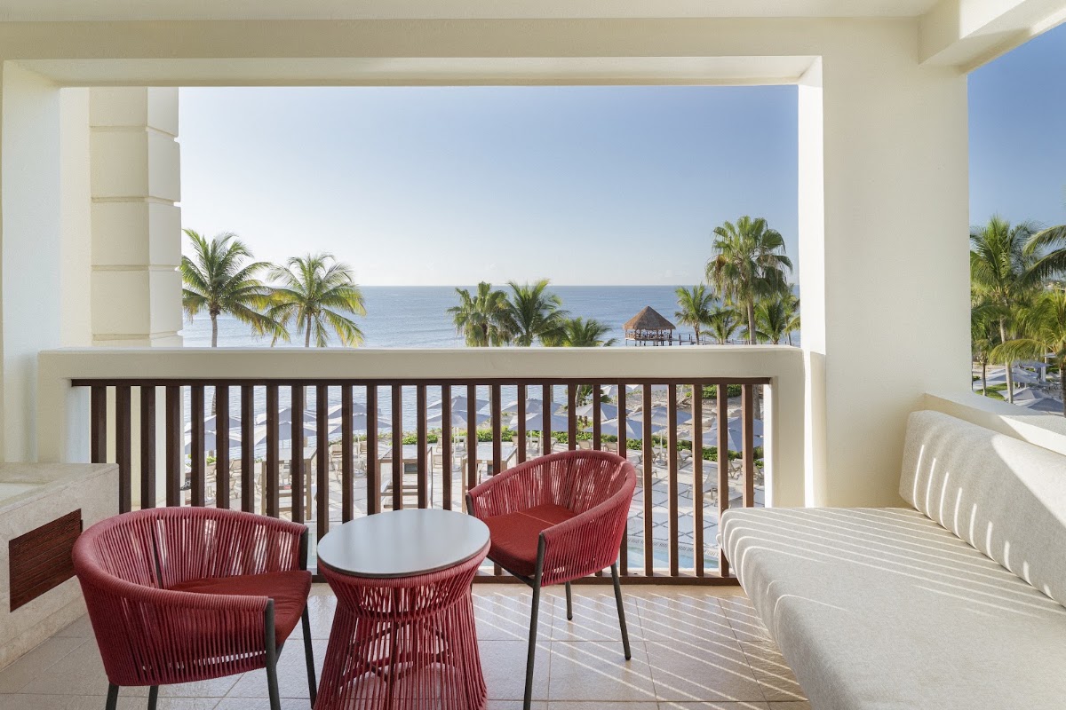 Oceanfront balcony with red woven chairs, small table, and views of palm trees and blue water at Dreams Puerto Morelos Resort & Spa.