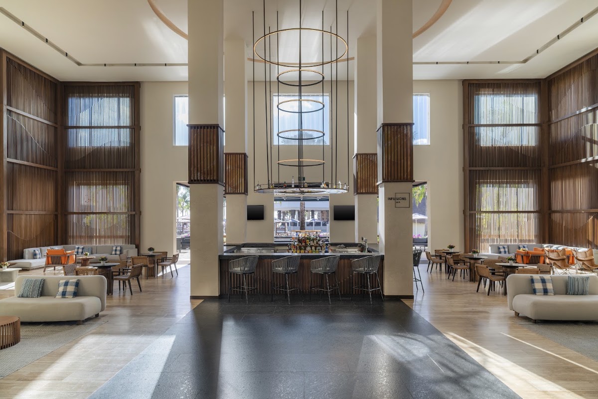 Modern lobby with high ceilings, central bar, geometric pendant lights, cream and wood tones, and seating areas with orange accents at Dreams Puerto Morelos.