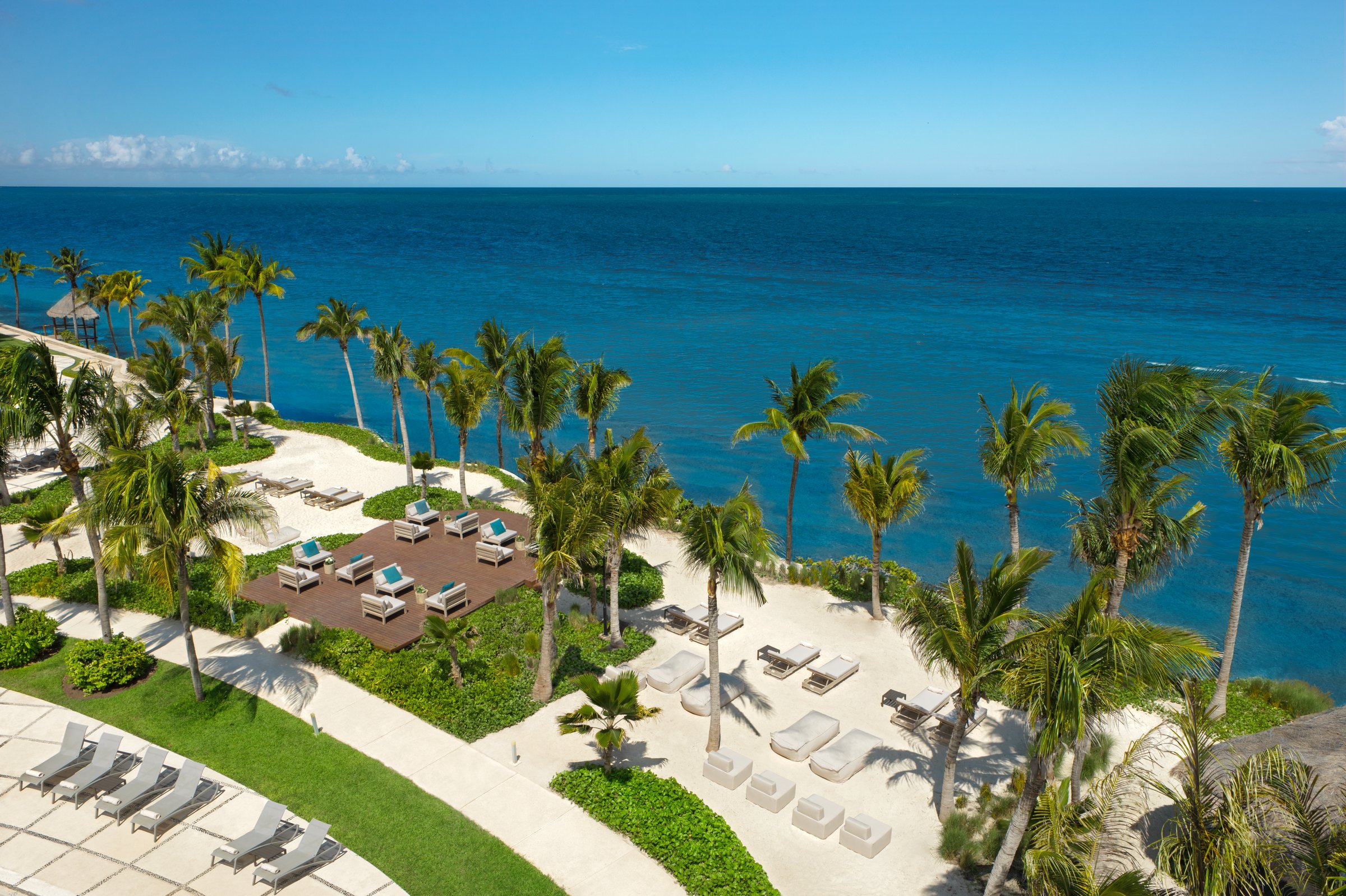 High view of the Papaya Sand lounge area, a dark wood deck with white and turquoise chairs and grey loungers on sand, beside the turquoise sea.