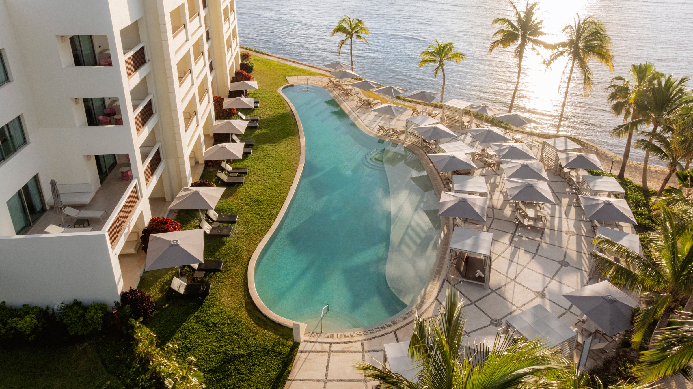 Aerial of the winding Preferred Club pool with grey umbrellas and cabanas on a patterned stone deck beside the palms and the sea.