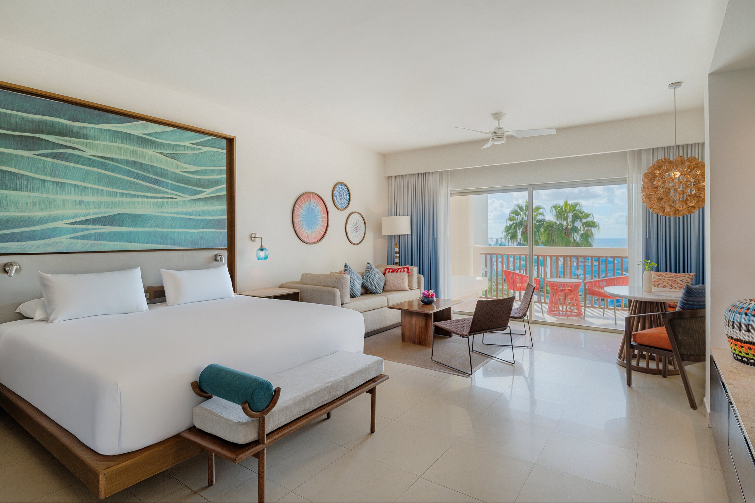 Junior suite with a king bed, a blue and green abstract headboard, a sofa area and sliding doors to a balcony with red chairs.