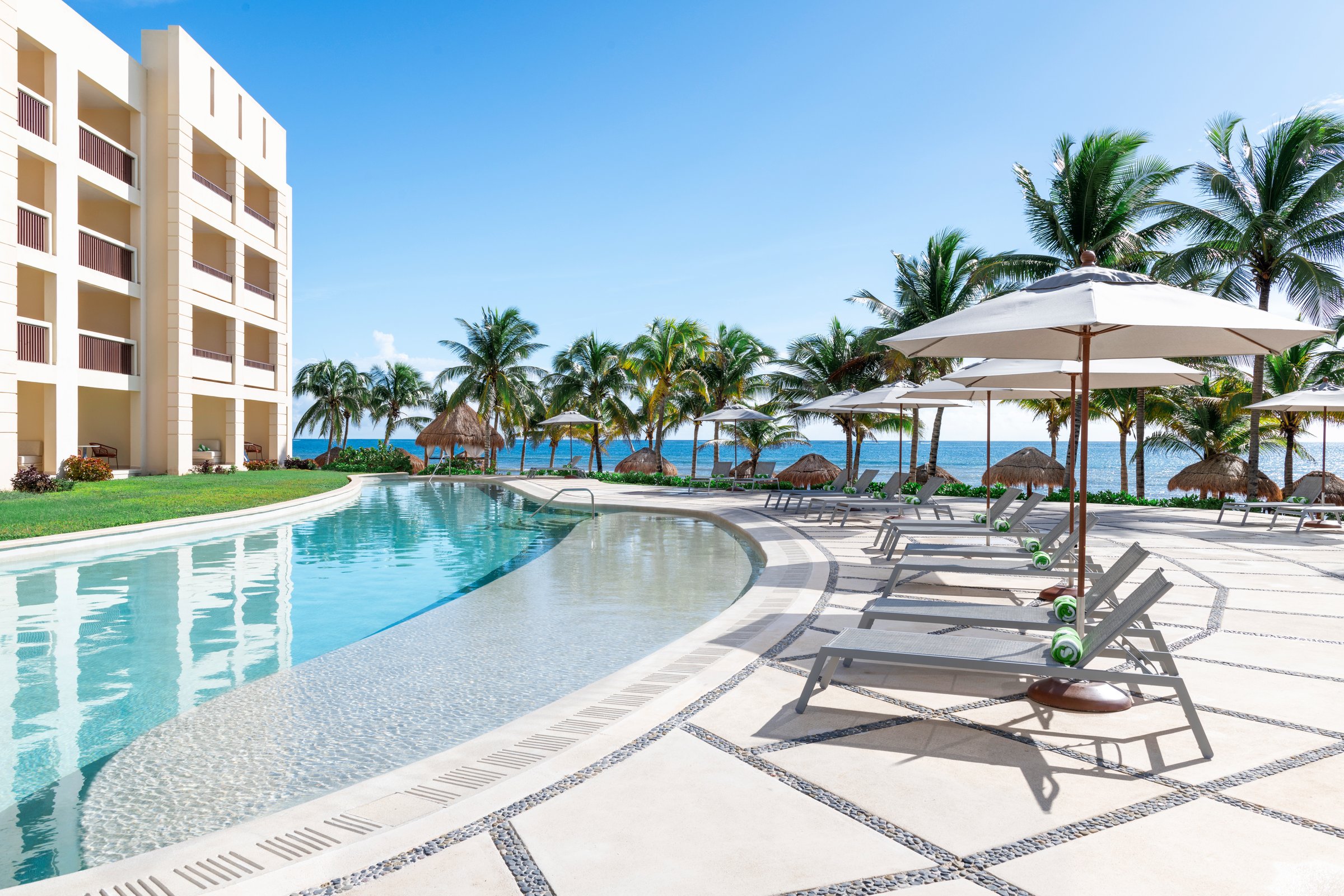 Curving infinity-edge pool with mosaic tiling and a stone patio of grey loungers and white umbrellas, with a white balconied building and palms.
