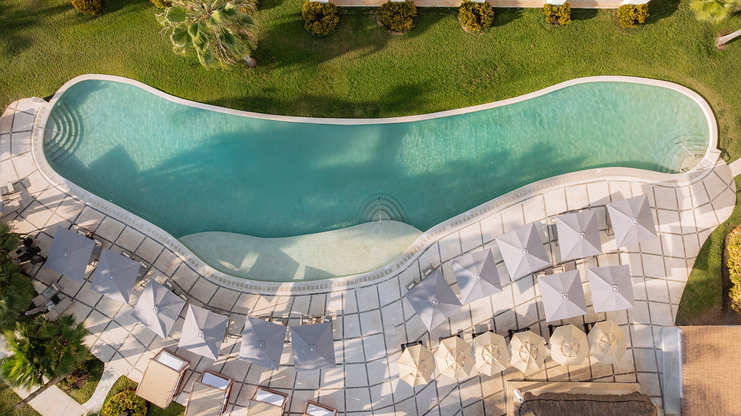 Top-down aerial of the wavy turquoise pool with a wide deck of grey umbrellas and loungers beside a green lawn.