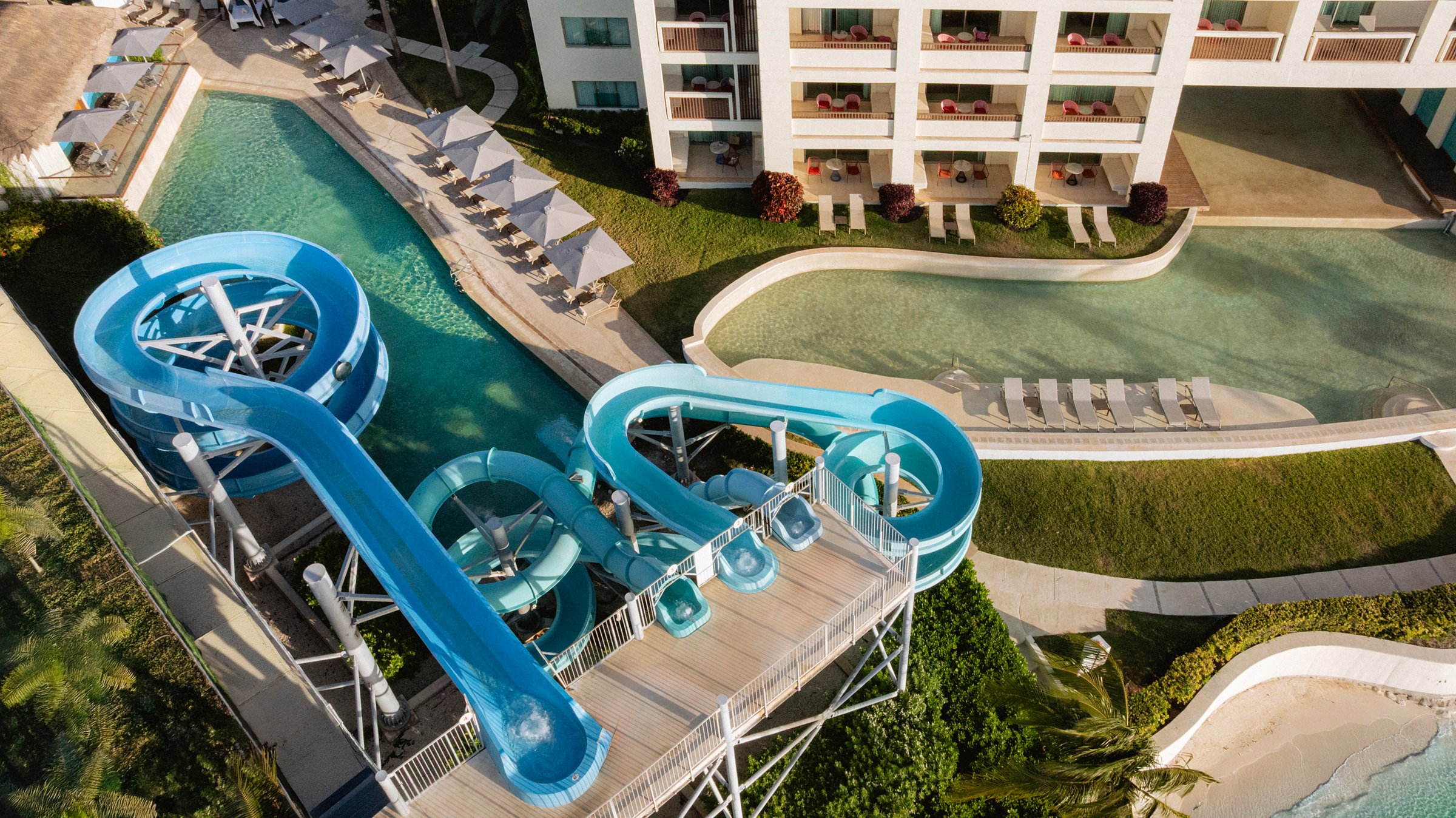 Aerial of the water park with two bright blue winding slides beside a lazy-river pool with loungers, framed by the white balconied resort building.