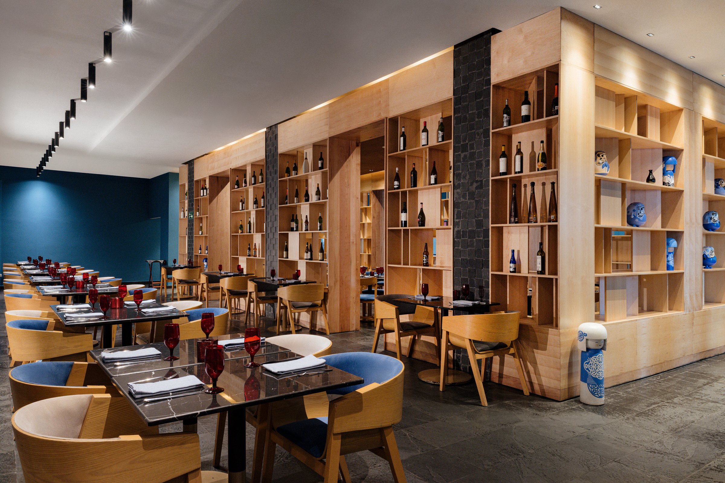Miyako dining room with light wood shelving of wine and sake bottles, small black tables and curved tan and blue chairs.