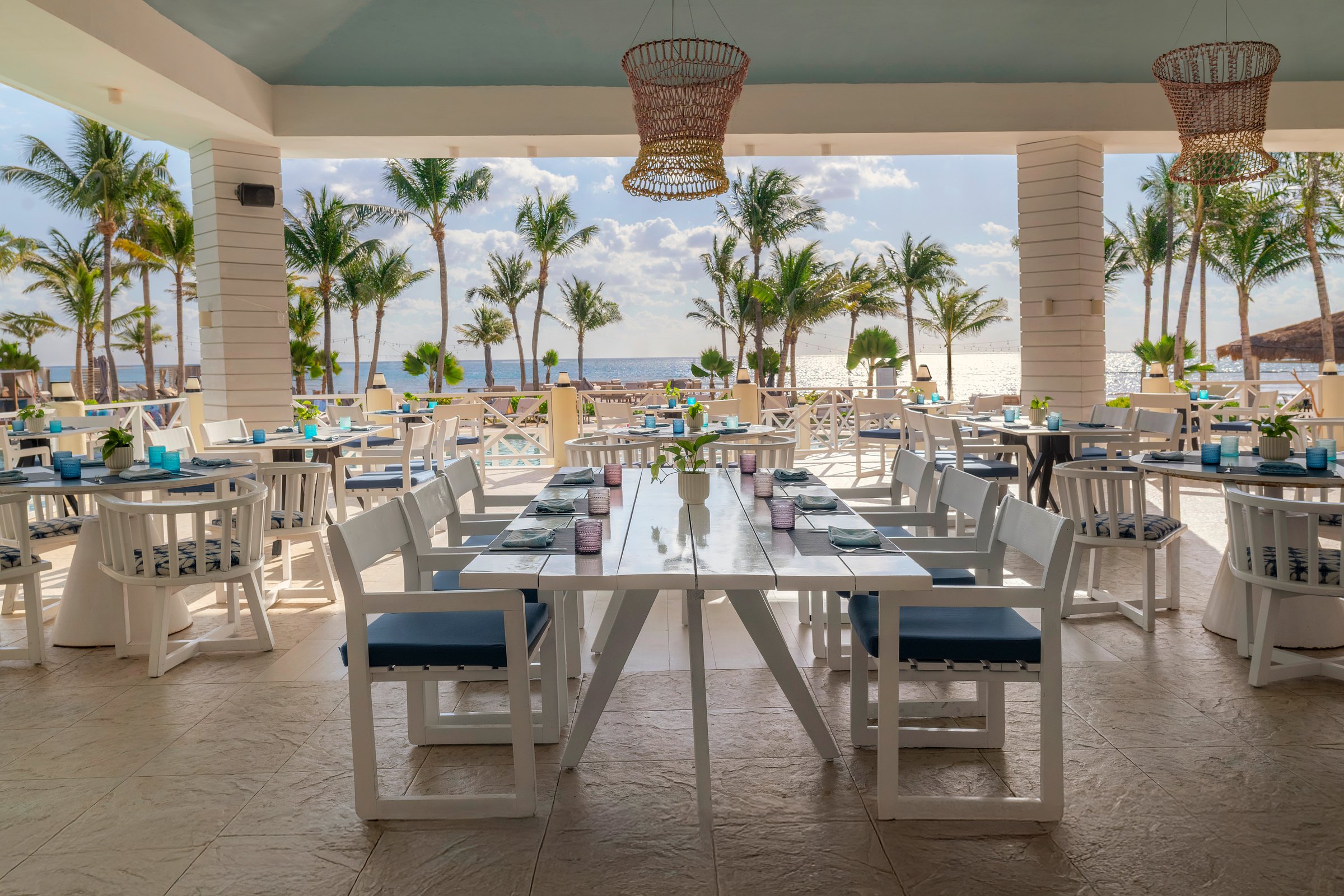 Balik open-sided dining pavilion with white tables, navy cushioned chairs and woven pendants, with palms and the sea behind.