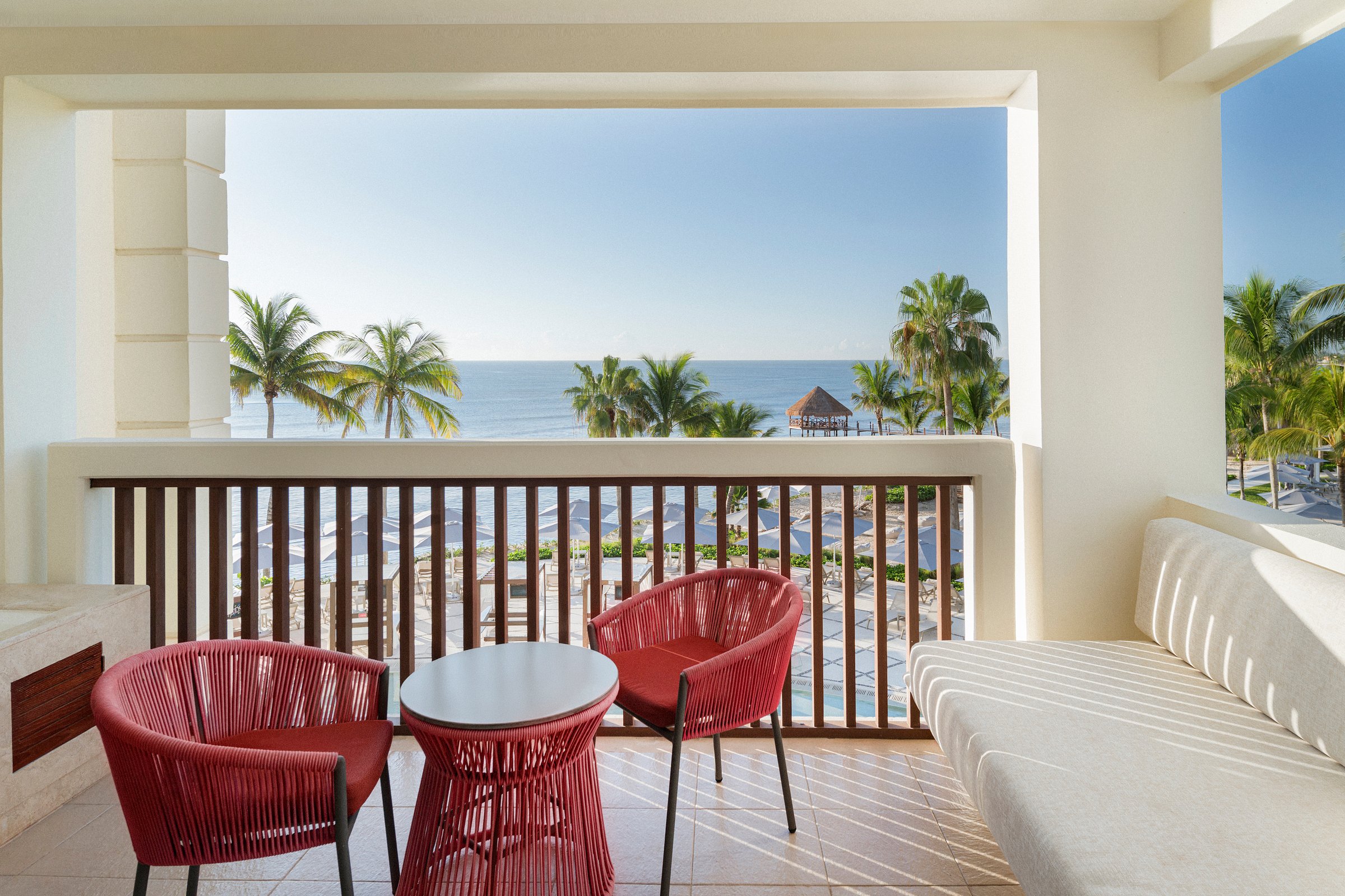 Balcony with two red woven chairs, a round table and a white sofa, looking over palms and a palapa to the turquoise sea.
