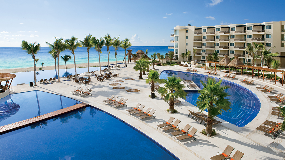 Dreams Riviera Cancun Resort & Spa pools and beach with palm trees, lounge chairs, and turquoise Caribbean Sea beyond.