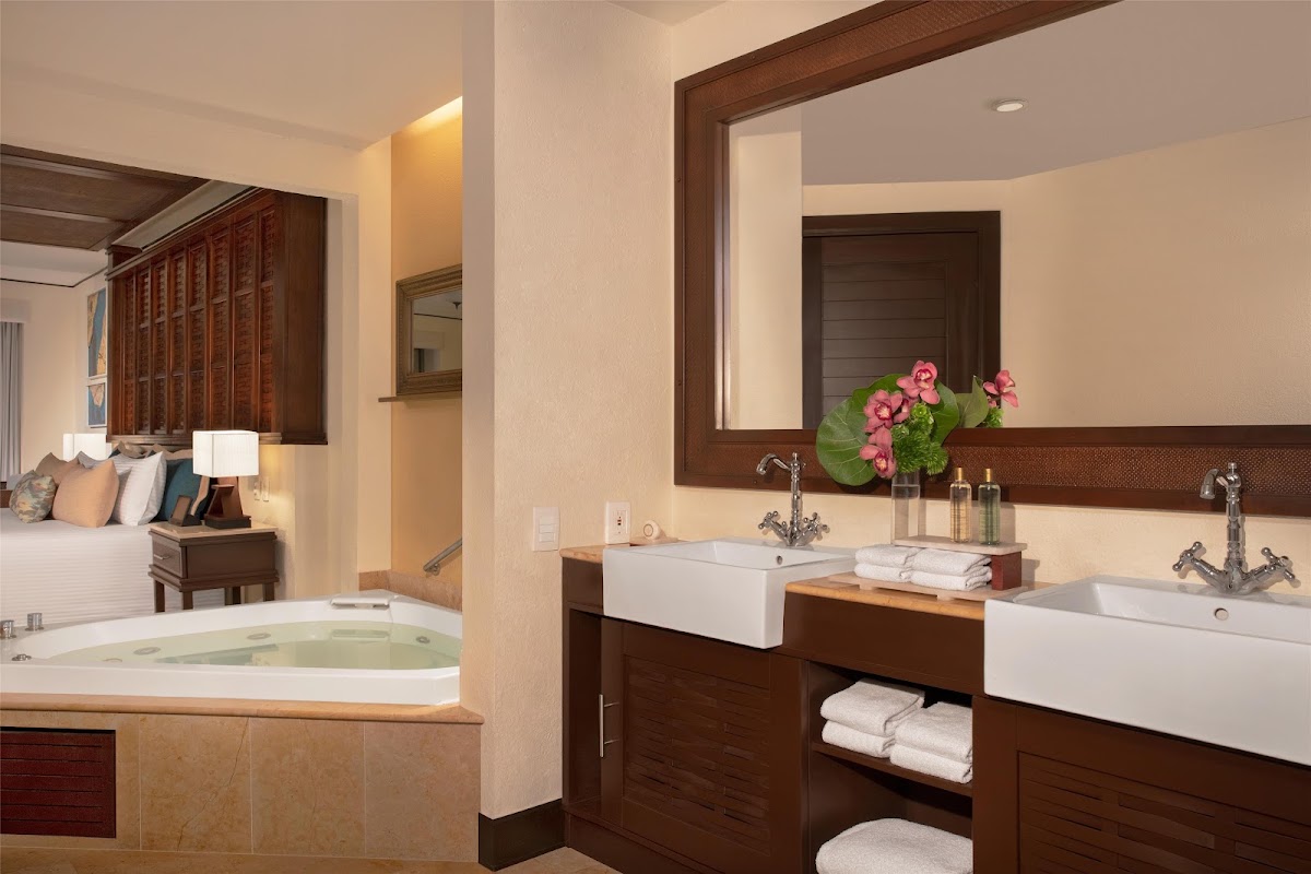Spa bathroom with soaking tub, double vanity sinks, and pink flowers, adjoining bedroom visible at Dreams Riviera Cancun.