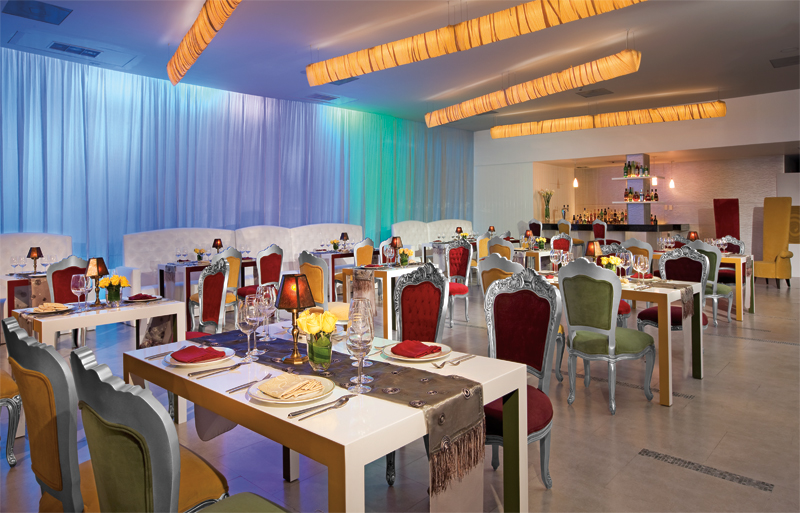 Dining room at Dreams Riviera Cancun with colorful chairs, set tables, warm ceiling lights, and blue-green curtain backdrop.