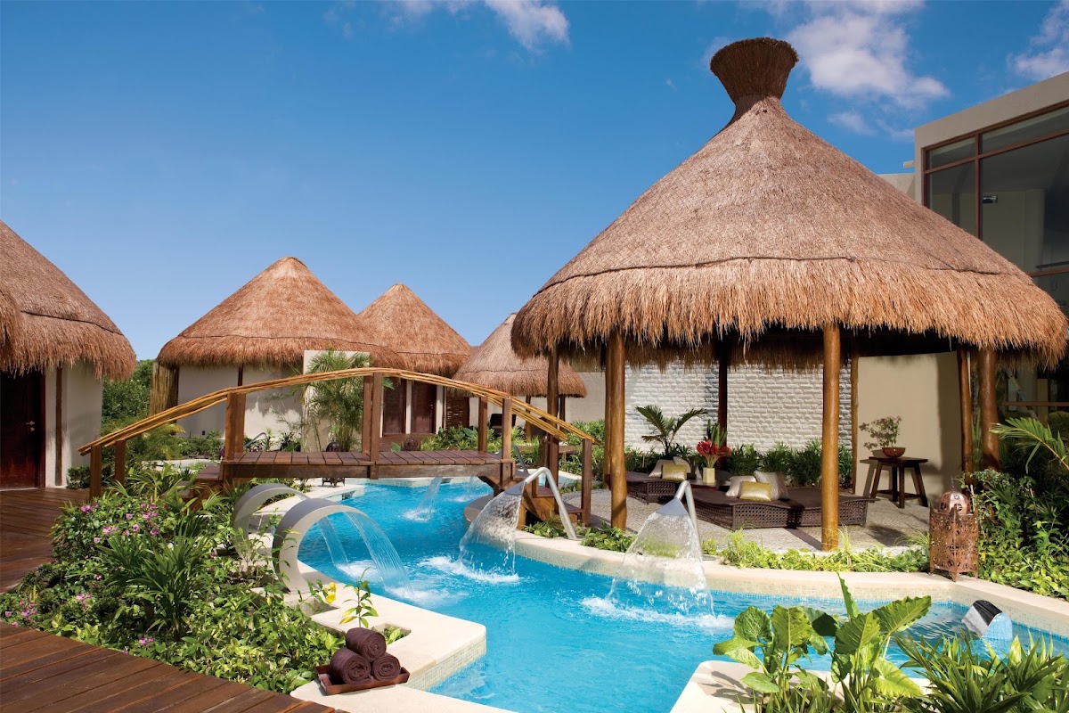 Thatched-roof villas with wooden bridges surround a blue pool with waterfall features at Dreams Riviera Cancun Resort & Spa.