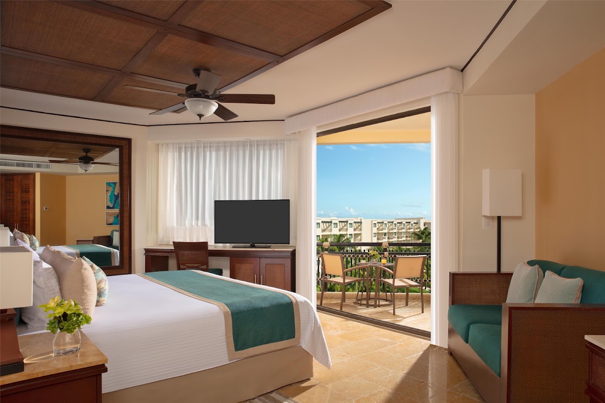Resort suite with king bed, teal runner, TV, and open sliding doors to balcony overlooking resort grounds and ocean at Dreams Riviera Cancun.