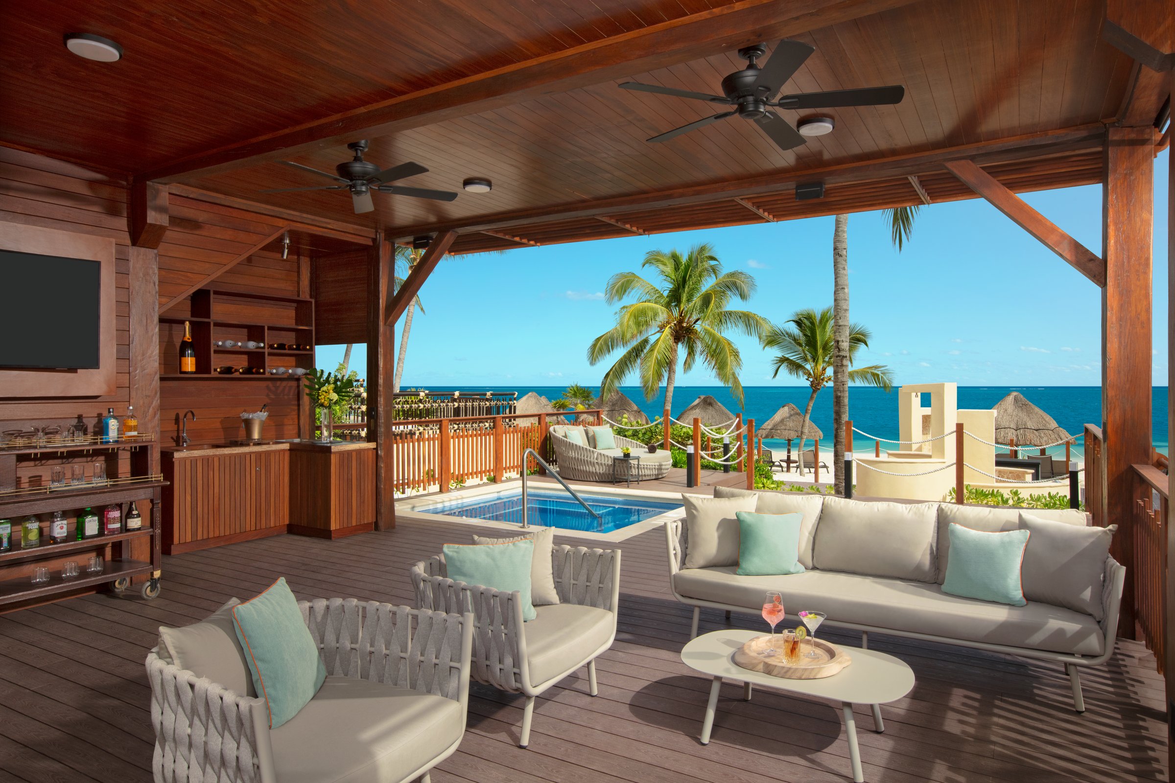 Shaded beachfront cabana with a grey wicker sofa and armchairs, a small plunge pool and palms, with the sand and sea beyond.