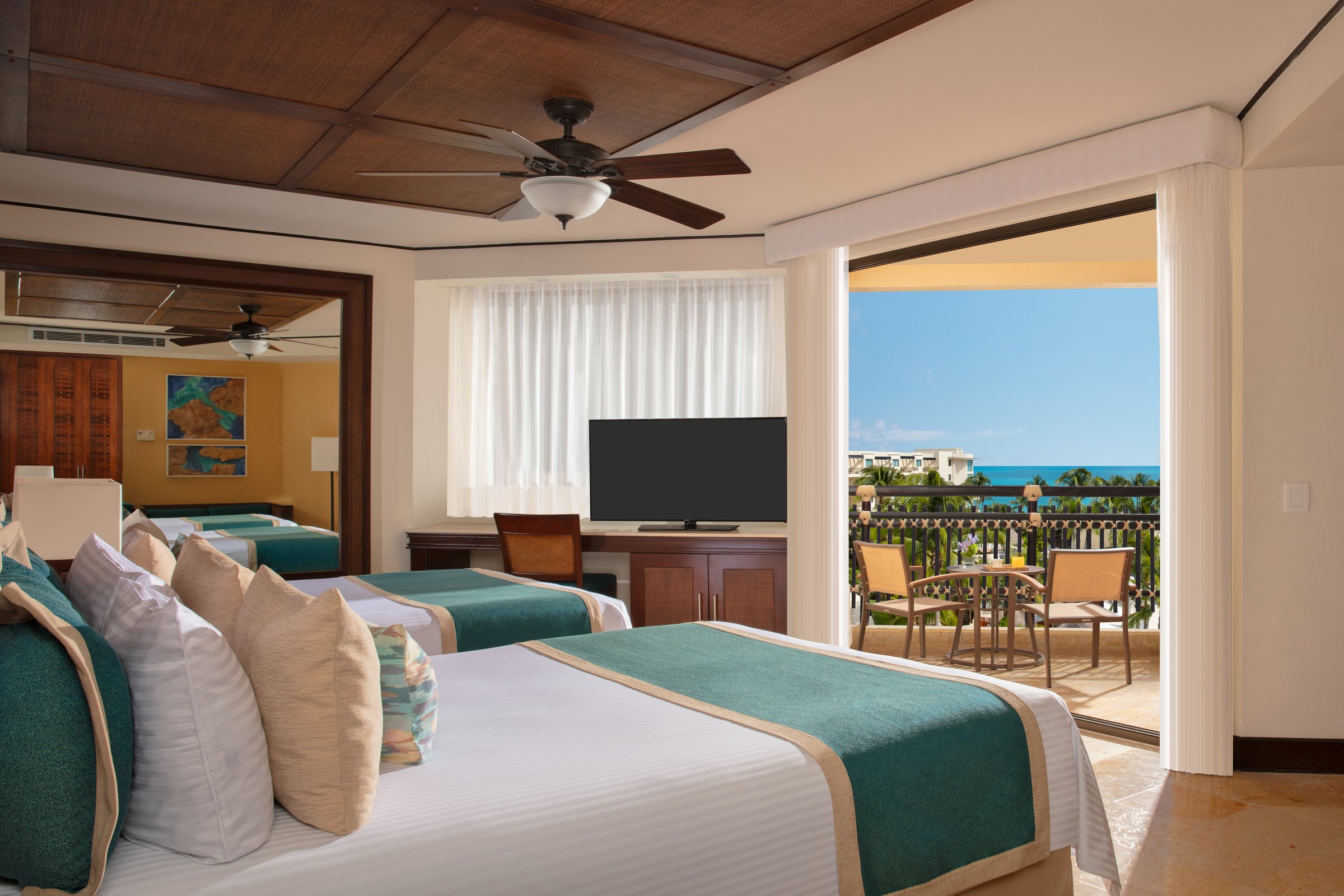 Deluxe ocean view room with two double beds and teal runners, with a balcony looking over palms to the turquoise sea.