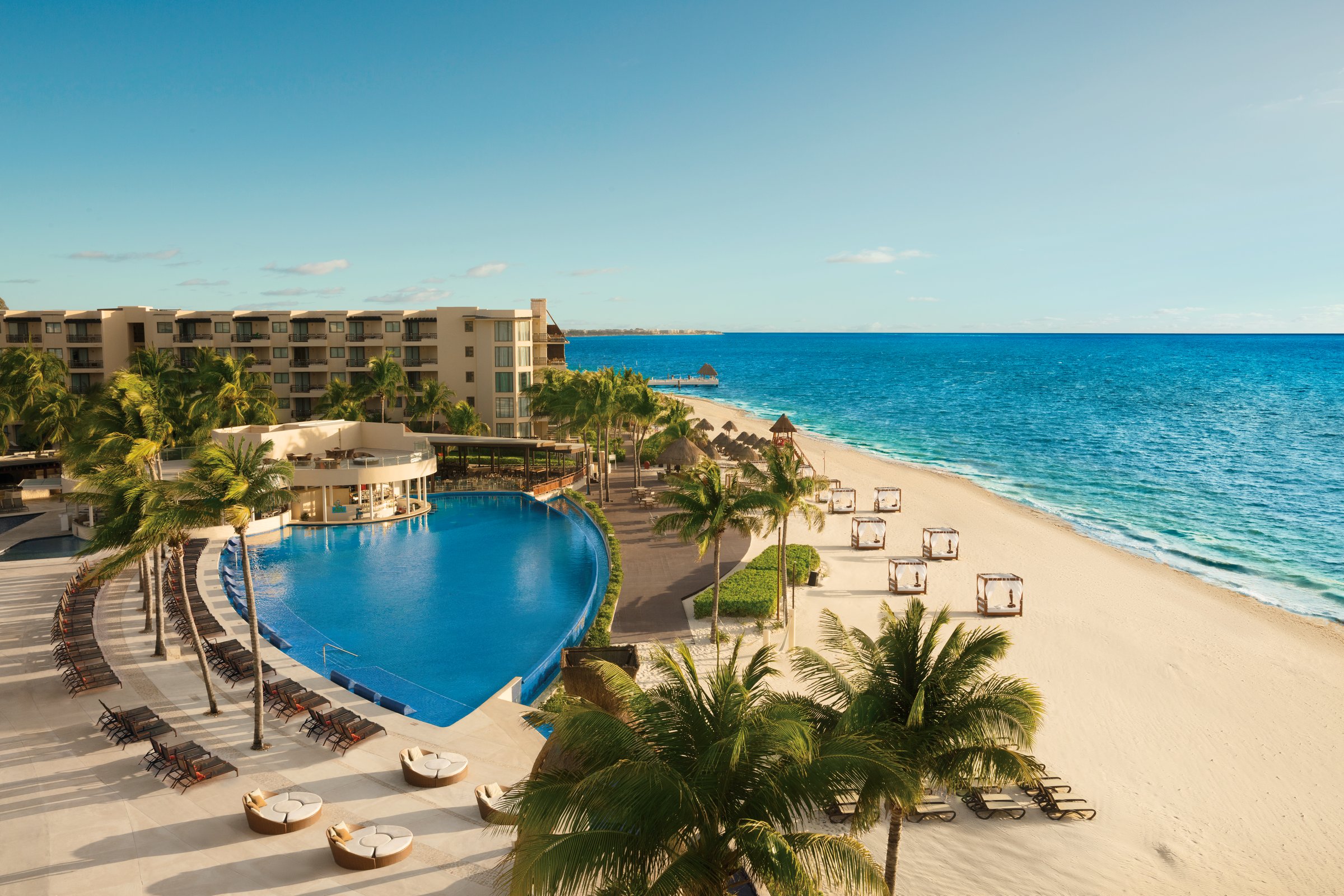 Elevated view of the curved main pool with lounge chairs and round daybeds beside a white beach lined with cabanas and a turquoise sea.