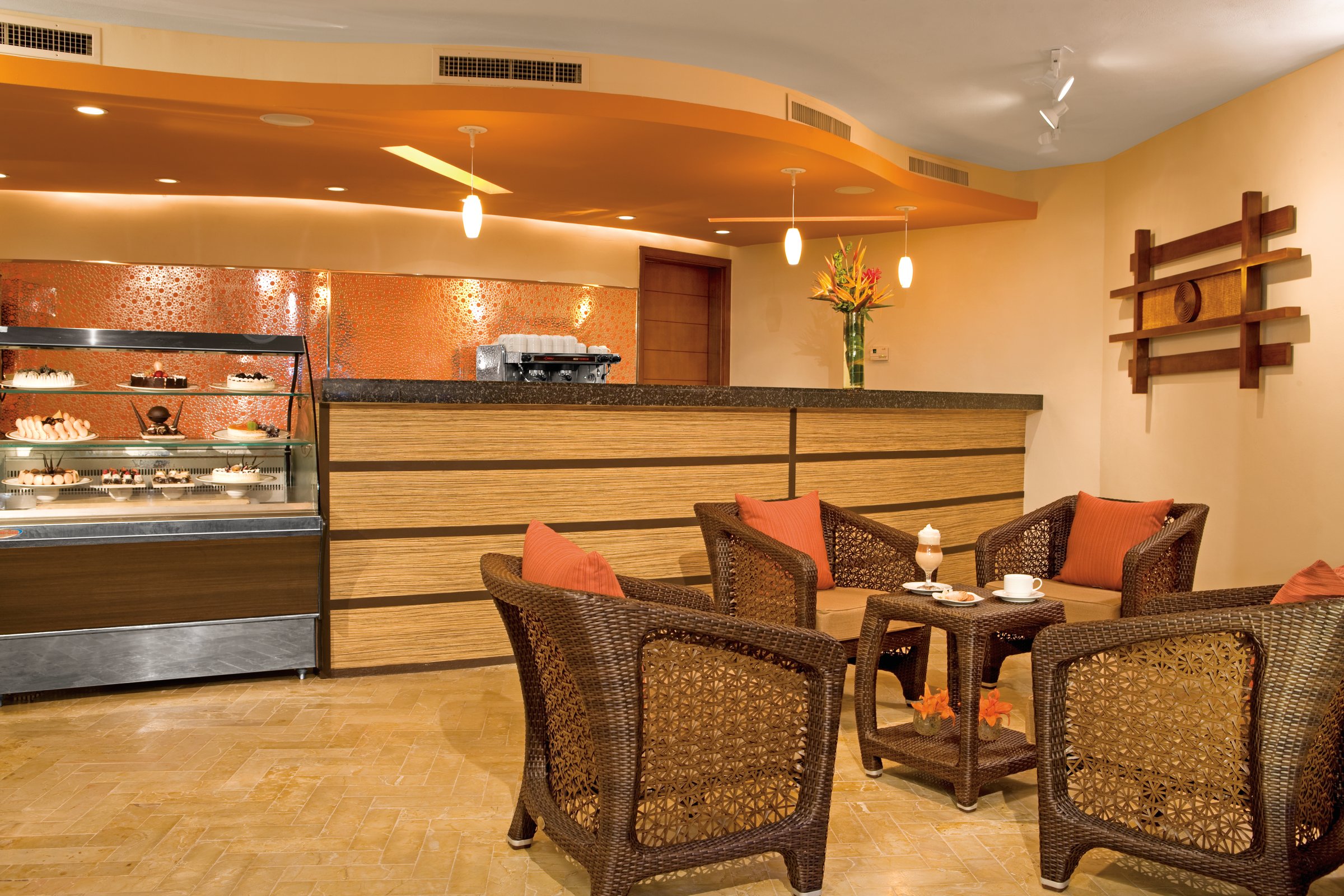 Coco Cafe seating area with wicker armchairs and orange cushions facing a counter with a glass case of cakes and pastries.