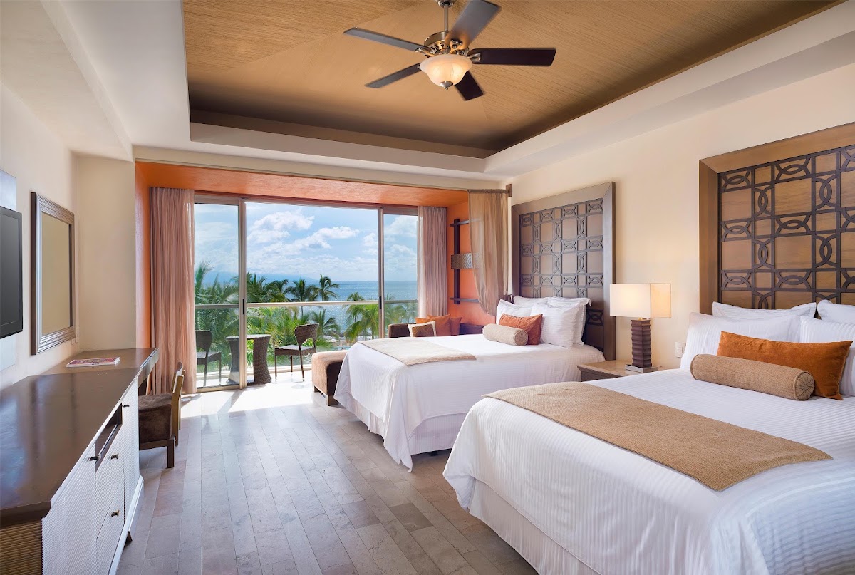 Spacious resort bedroom with two queen beds, ocean and palm tree views through sliding glass doors, and ceiling fan overhead.