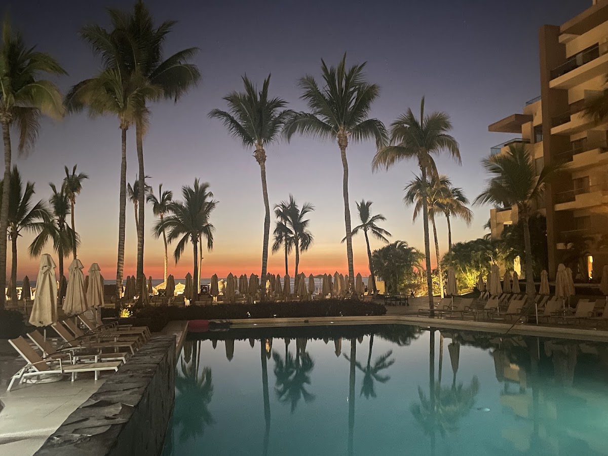 Resort pool at dusk with palm trees reflected in still water, lounge chairs and umbrellas lining the deck, and a warm sunset sky beyond.